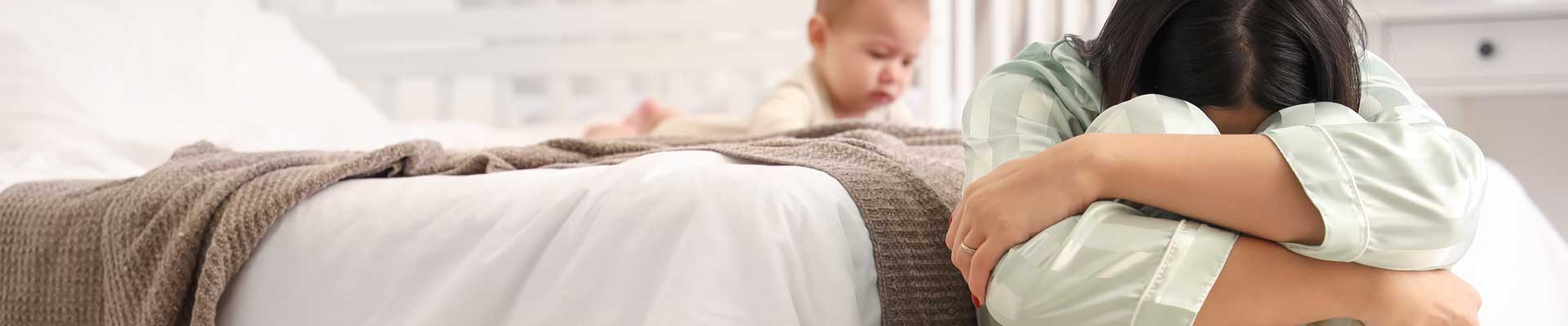 a young mother experiencing mental health difficulties expressed with a young woman sitting on the floor with her head down next an infant crawling on the family bed beside her