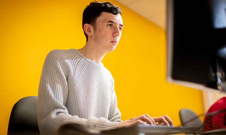 Male student sat against a yellow wall working on a computer 