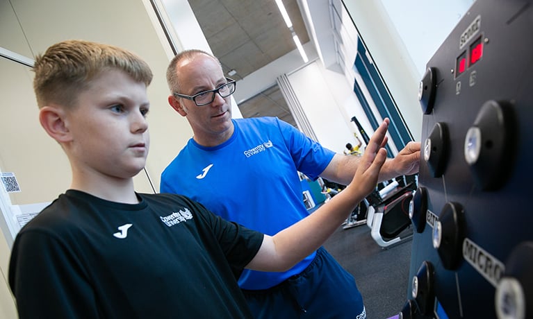 A postgraduate student assisting a school student with an exercise machine.