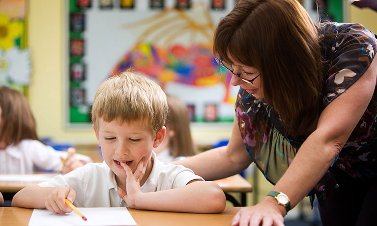A female teacher helping a pupil with his work as he sits at a desk with a sheet of paper in front of him, holding a pencil.