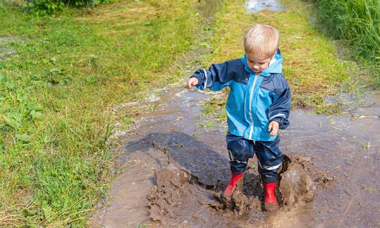 a toddler wearing blue coat and boots is jumping in a muddy puddle of water on a field