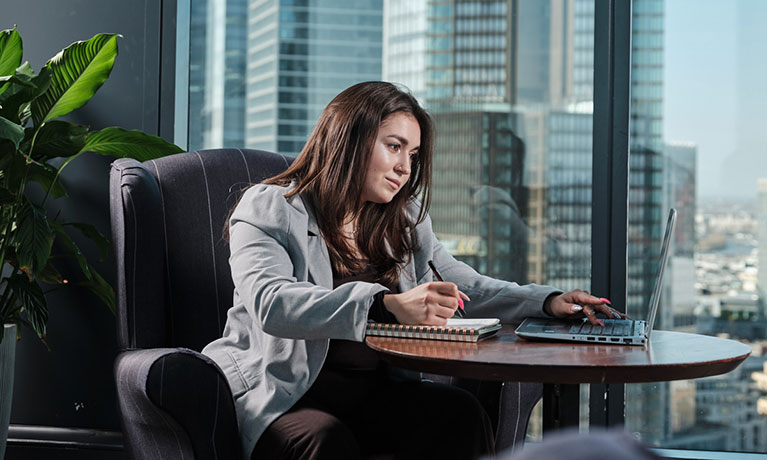 A student working in front of a large window with the London skyline behind her.