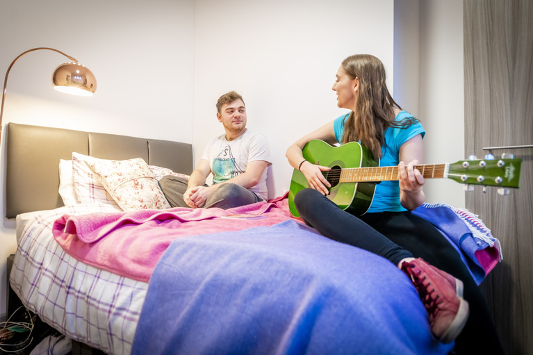 Two students on a bed in student accomodation one playing a guitar