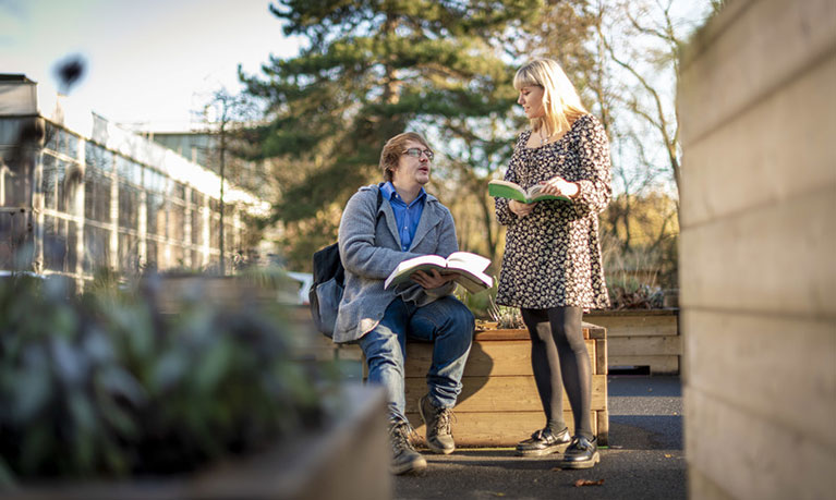 Male and female students sitting outside on campus holding books 