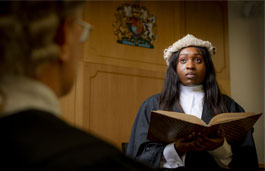 Female student standing in a mock court room wearing  robes and wig, holding an open book