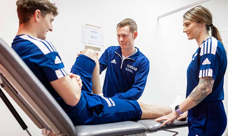 A student treating a student on a hospital bed with another student looking on. 