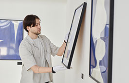 A man wearing white gloves adjusts the hanging of a painting in a gallery
