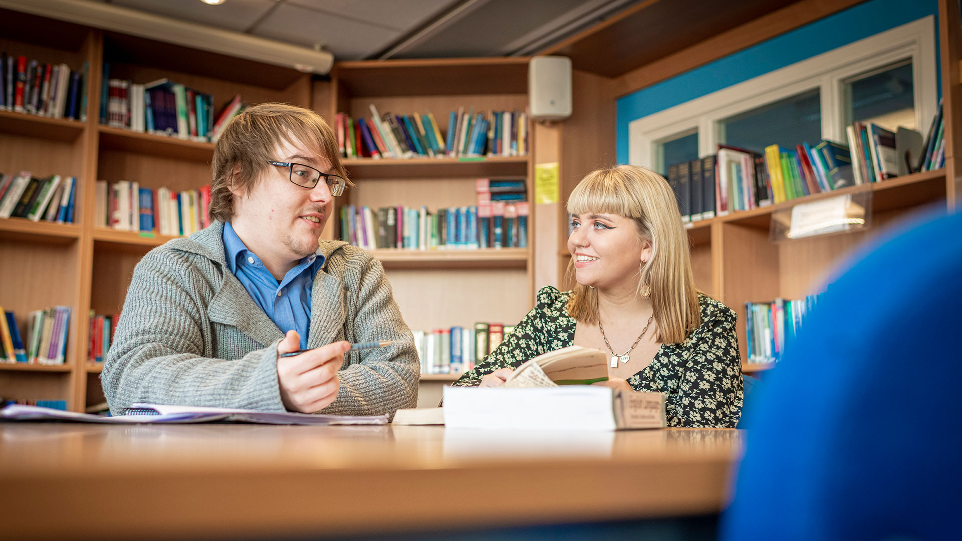 Two students sitting at a desk, talking, with bookshelves behind them