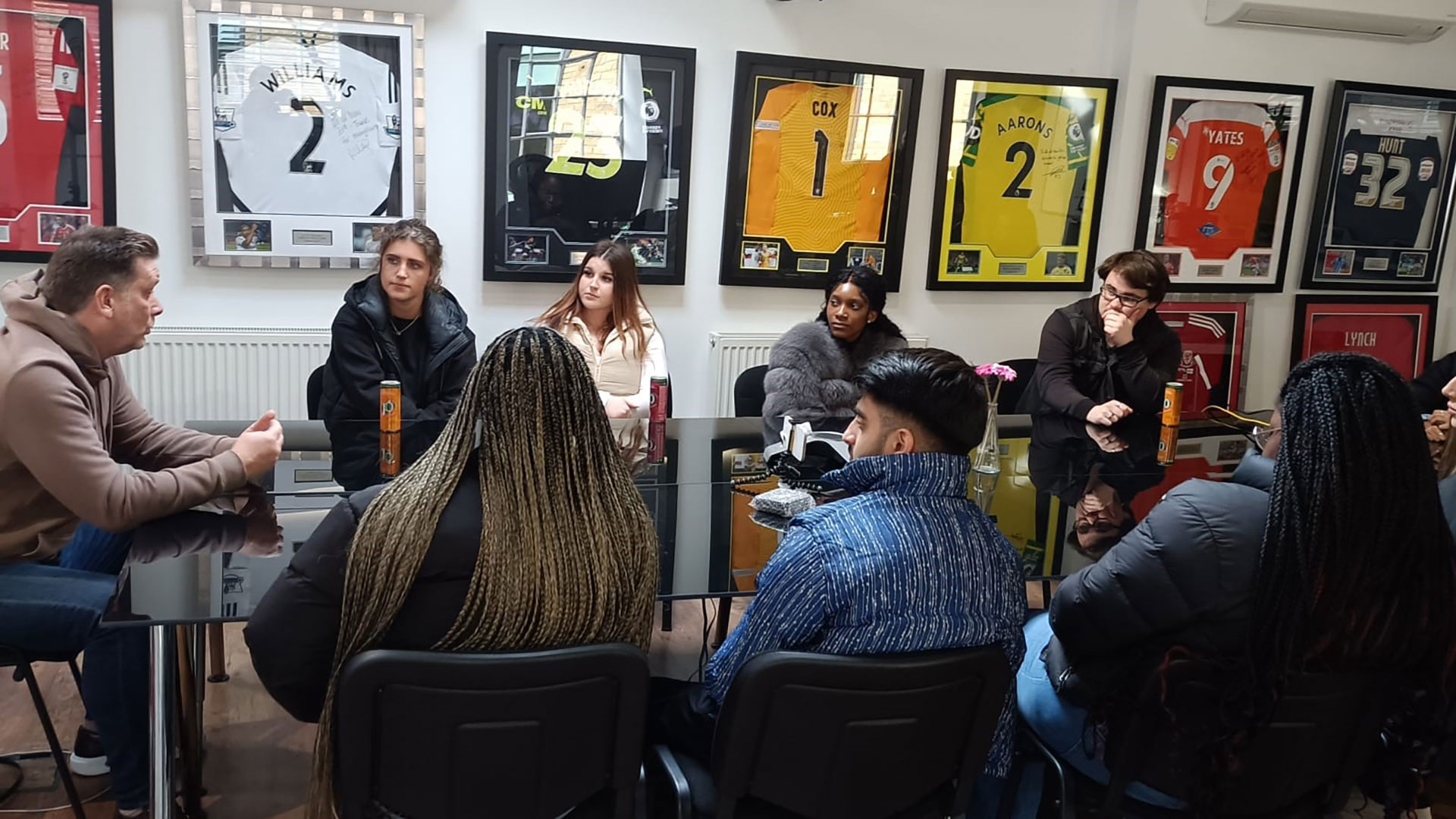 Lecturer and group of students at a boardroom style table with lots of football shirts behind them