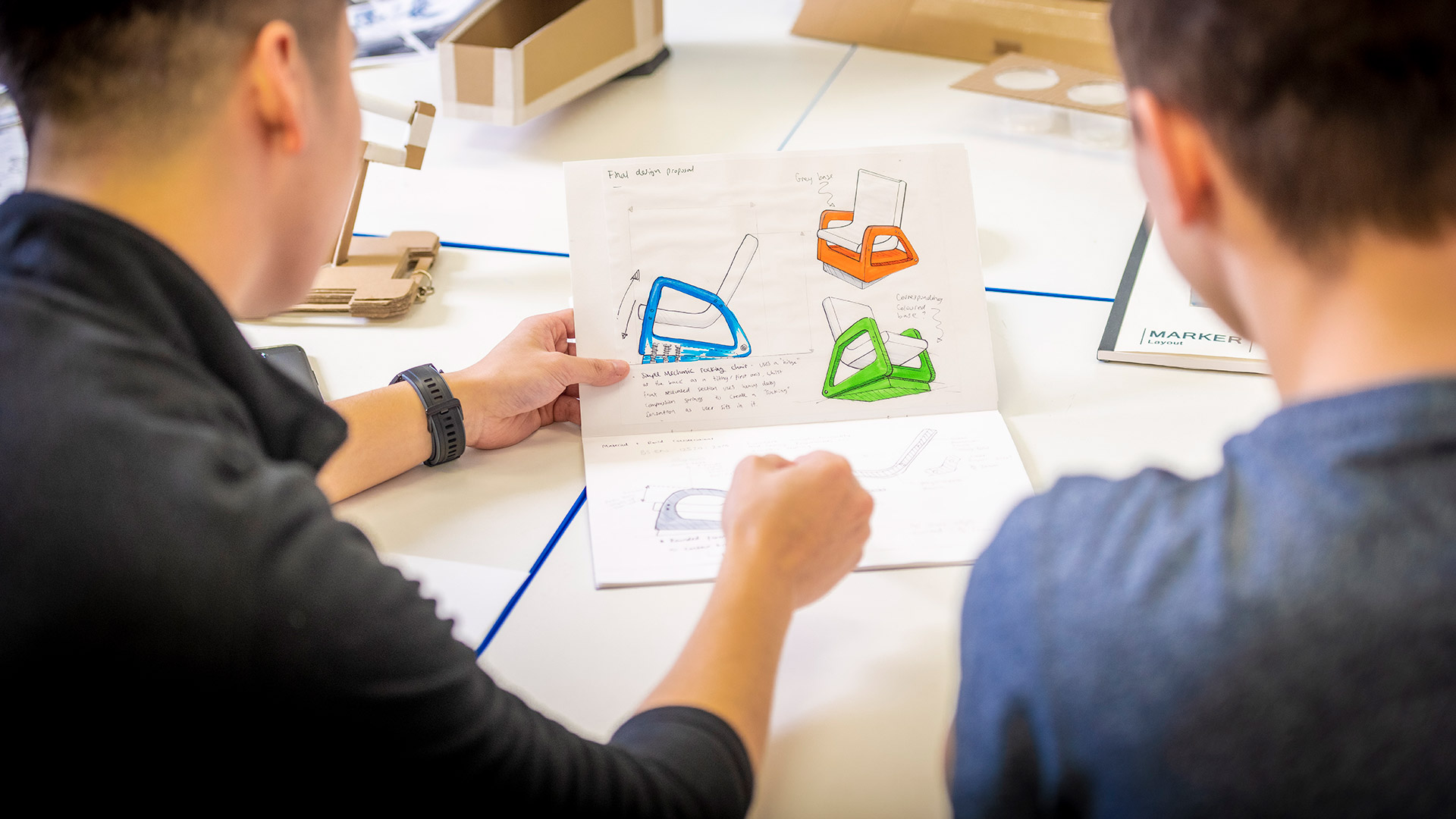 two male students looking at colourful product designs on a design sheet 