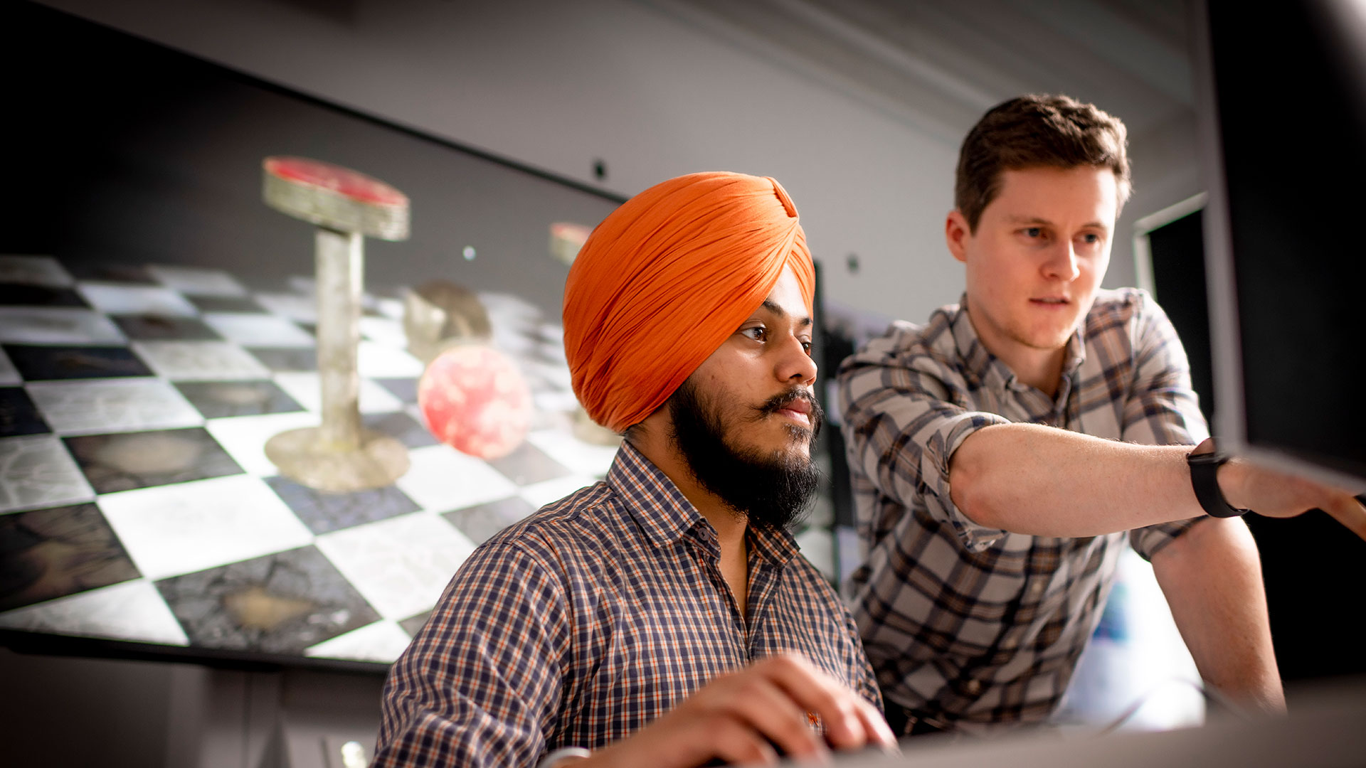 Student wearing turban looking at screen, tutor leaning over pointing at the screen