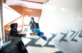 four smartly dressed people in front of a bright window face each other across a desk, one is standing