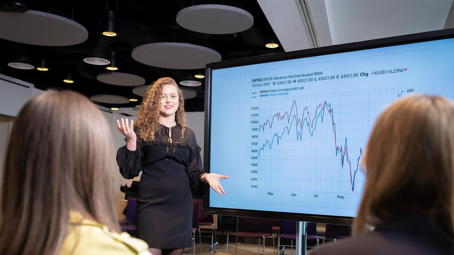 Woman standing in front of large screen, talking, while two people look at her