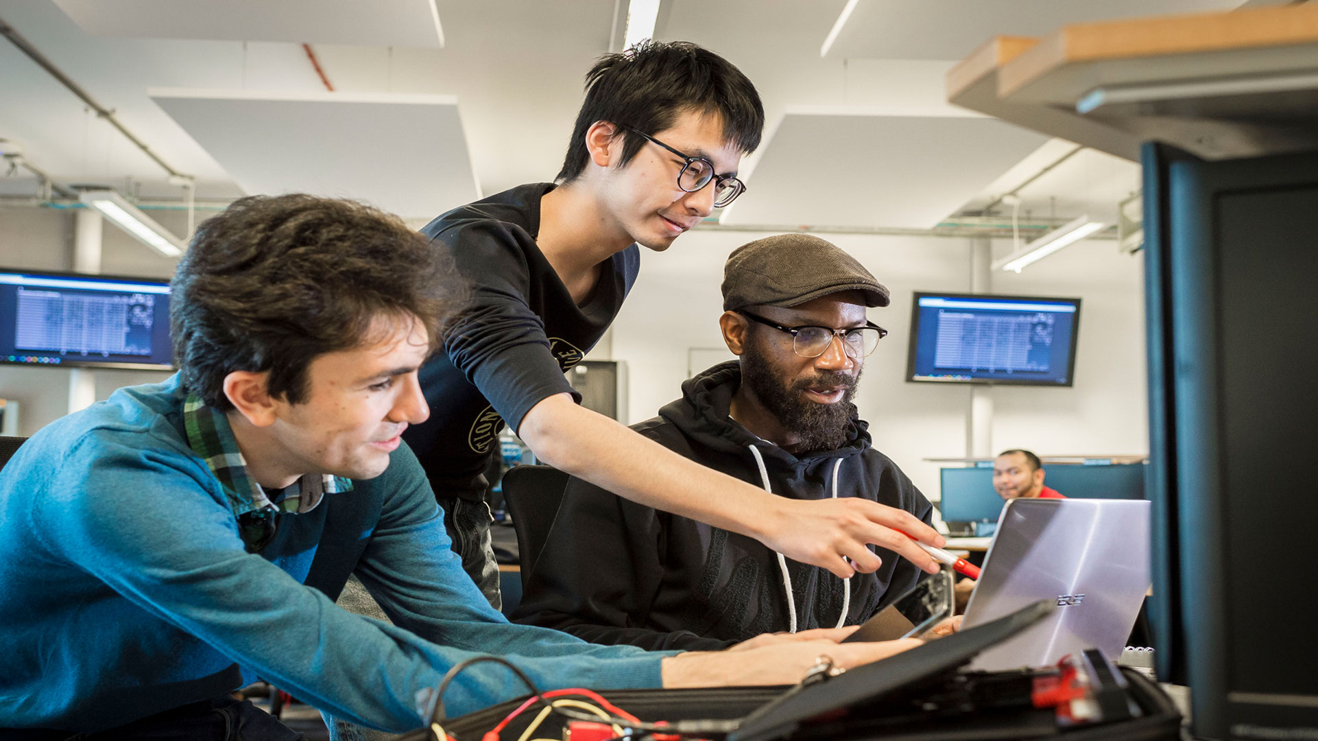 Three people lean over a screen, the middle one standing and pointing at the screen