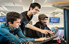 Three people lean over a screen, the middle one standing and pointing at the screen