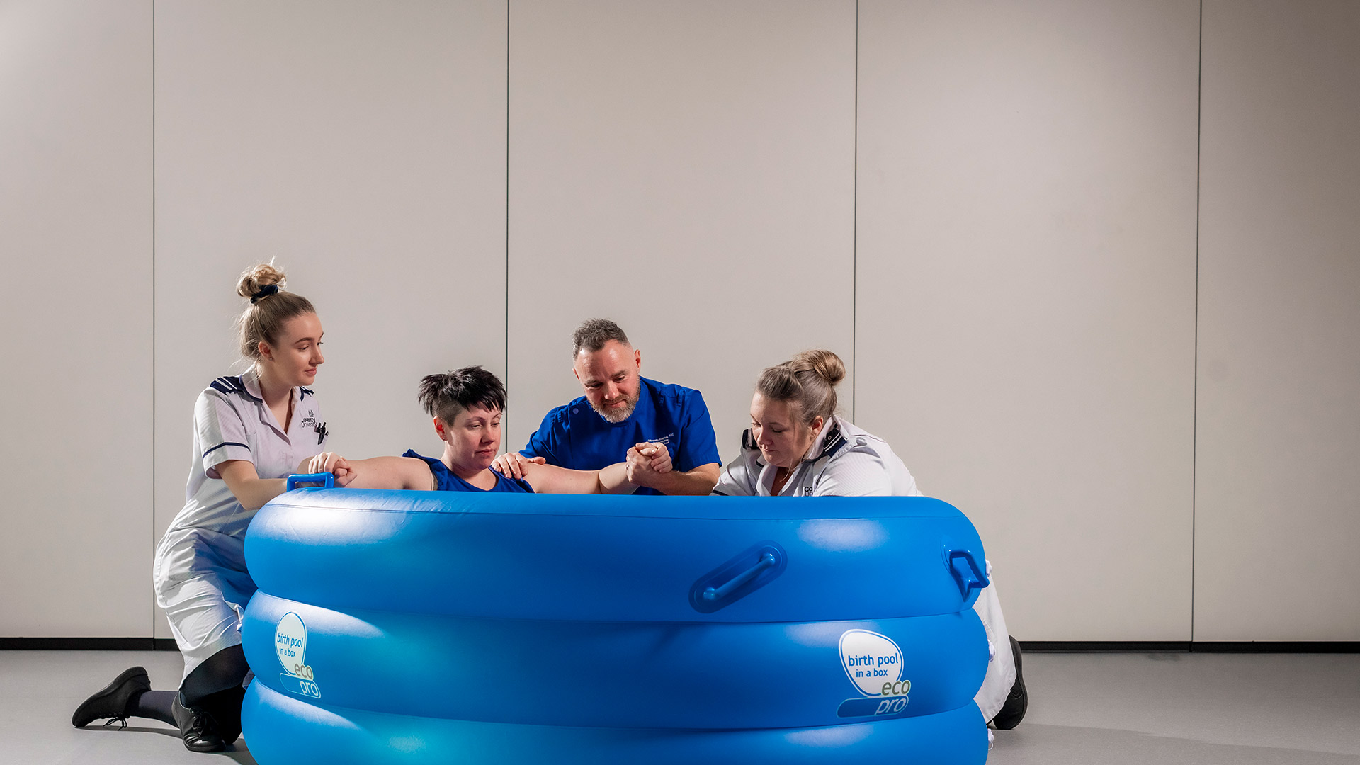 A woman giving birth in a blue water bath with two nurses either side and a visitor behind her for support