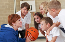 A teacher holding a basketball in a school sports hall, giving a team talk to a group of primary school-aged children.