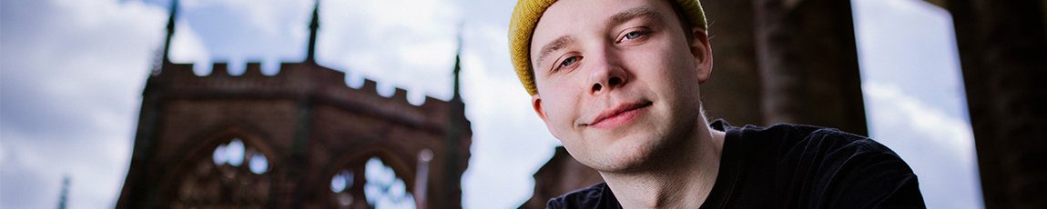 A student in a yellow woolly hat, smiling at the camera in the ruins of Coventry Cathedral.