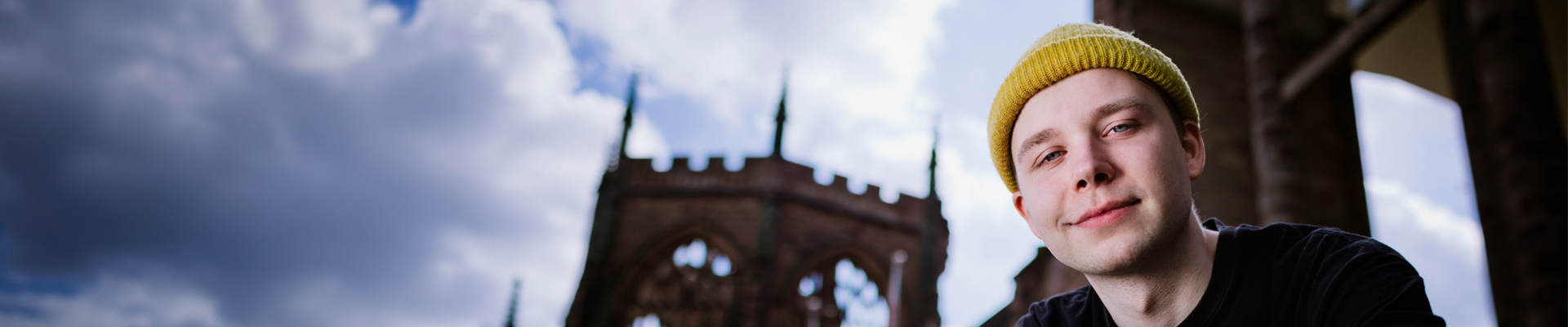 A student in a yellow hat looking at the camera with the Coventry Cathedral spire and ruins behind him.