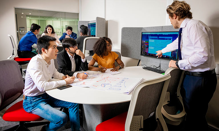 Three students sat at a desk while lecturer points at screen.