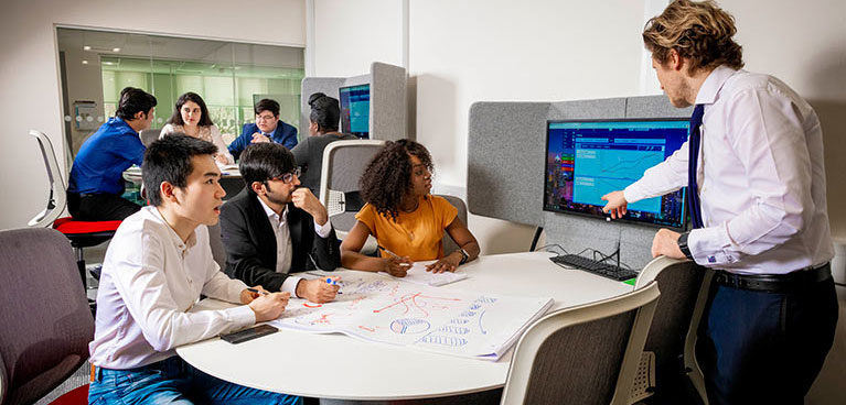 Three students sat at a desk while lecturer points at screen.