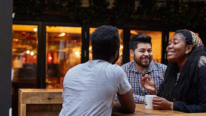 Three students sat outside in a coffee shop talking and laughing