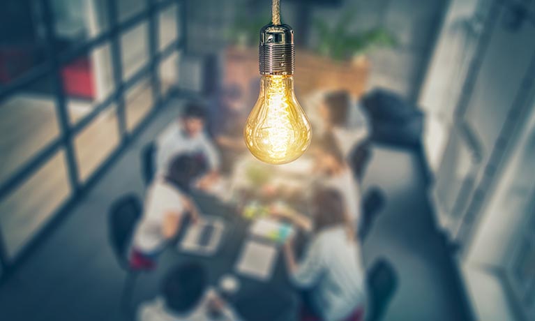 Lightbulb in foreground with people at meeting table below.