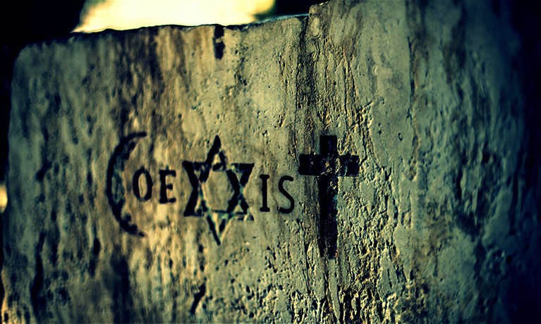 The word Coexist carved into a stone wall. Some of the letters are formed by the Islamic crescent, Star of David, and Christian Cross