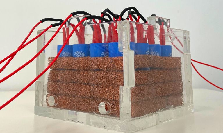 A close up of a Perspex box with 4 layers of copper coil, blue batteries and red cables 