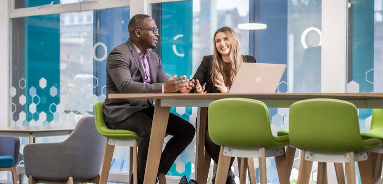 Male and female staff member chat at desk with laptop.