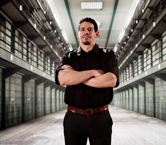 Person in a black uniform standing with arms crossed in a multi-level prison corridor.