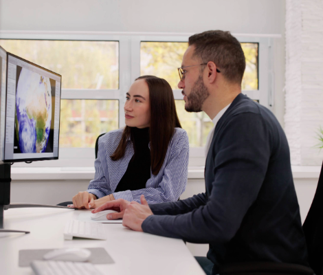 Two people sitting at a desk looking at a computer monitor displaying a world map.