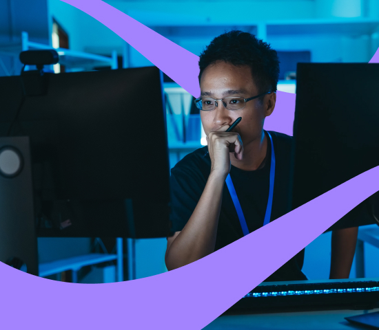 Person working at a desk with two monitors in a dim, blue-lit room, looking focused while holding a pen to their face.
