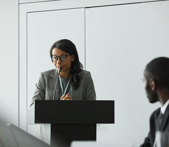 Person standing behind a podium speaking, with another person seated and facing them in a plain room.