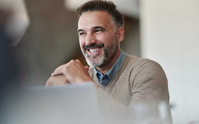 Smiling man with a greying beard sitting in front of a laptop 