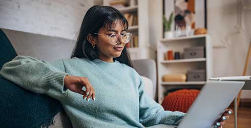Young female with long dark hair wearing glasses sitting on a sofa with a laptop on her lap