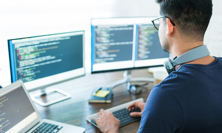 A programmer coding at a desk with two screens and a laptop open, wearing headphones around his neck.