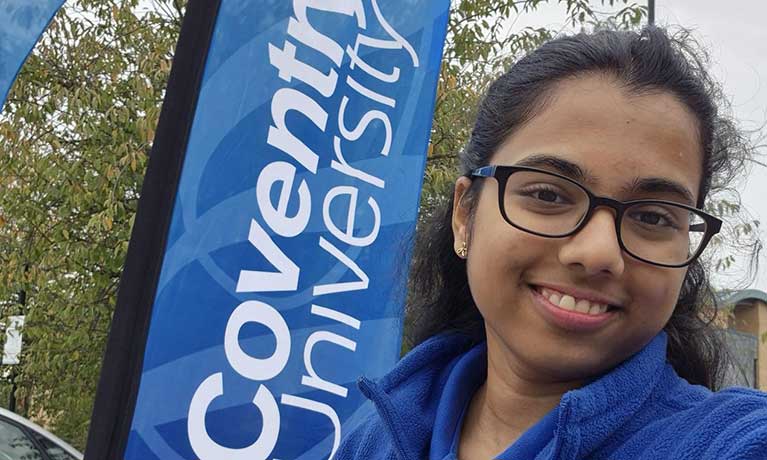 Sri standing outside the campus with the Cov University flag in the background Sri Anshula Chennareddy standing in front of a university banner