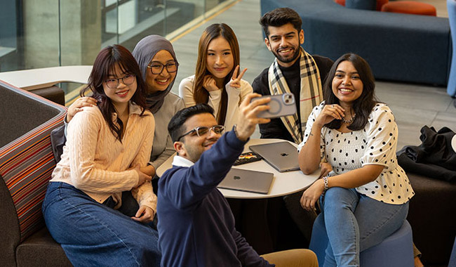 A group of students smiling around a table as one of them takes a selfie of them all with his phone.