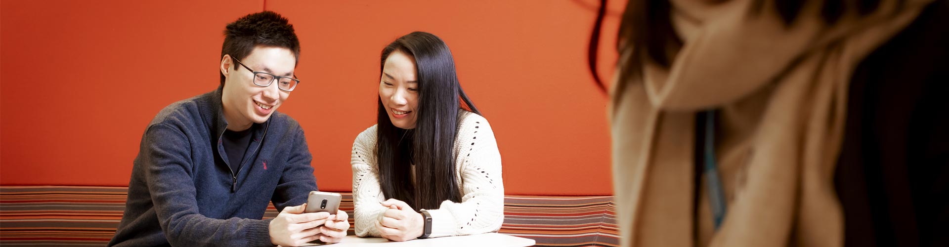 Two students smiling and looking at a smartphone in a study area on campus.