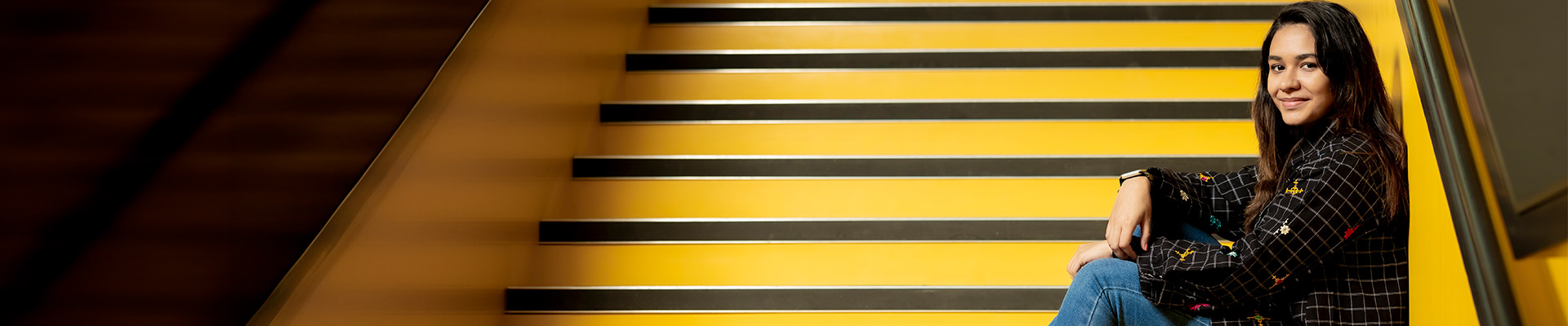 An international student sat on bright yellow steps in The Hub, wearing jeans and trainers and smiling at the camera.