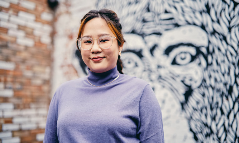 Someone looking at the camera smiling with a brick wall and mural in the background