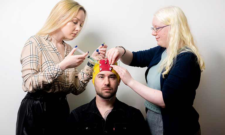 Two standing members of staff look at hat fitted to a man sitting between them