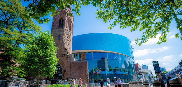 The Wave sports centre and Spire Bar in front of a backdrop of blue sky and surrounded by trees and people walking.