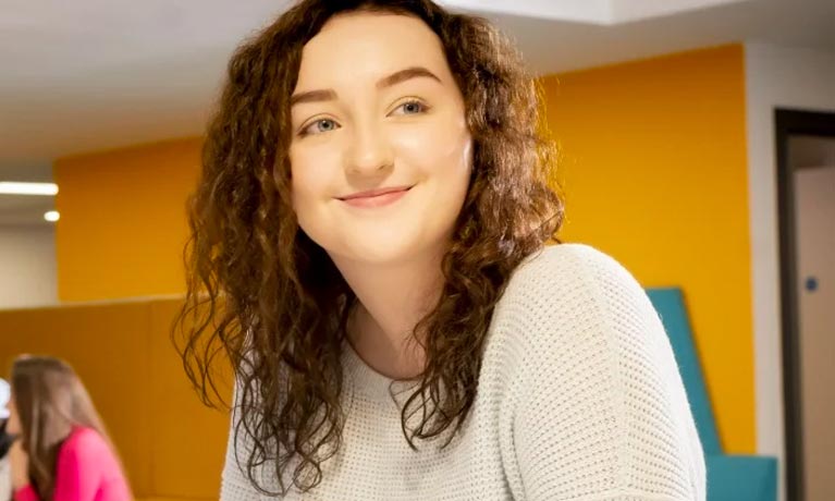 Female student sat against a yellow wall smiling