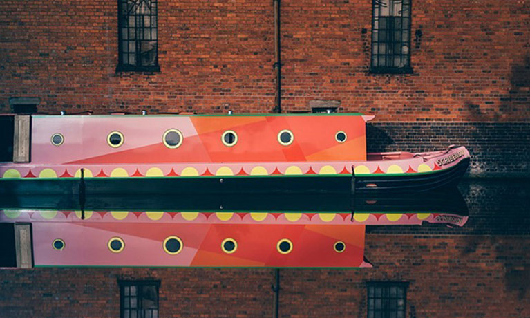 A canal boat moored up against an old warehouse building, reflecting on the water