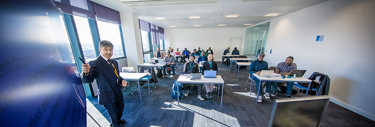 lecturer standing at the front of a classroom pointing at a screen and speaking to students