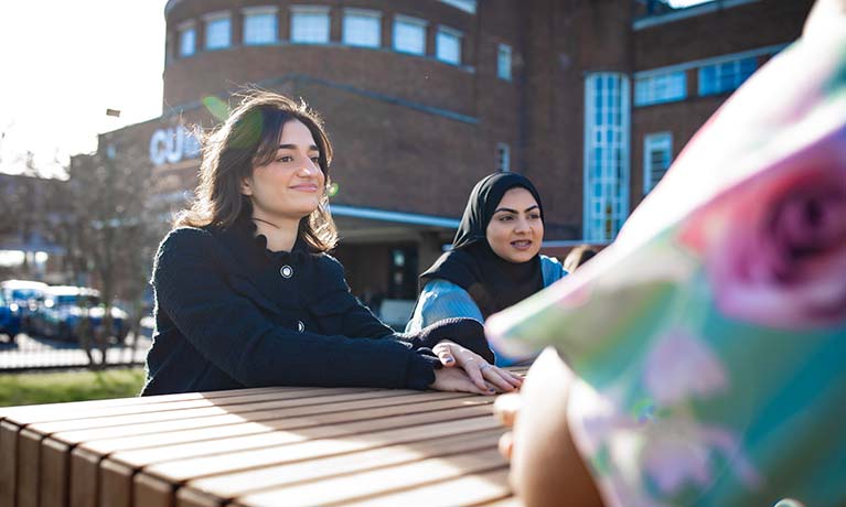 Two students sat on a bench outside, facing another student