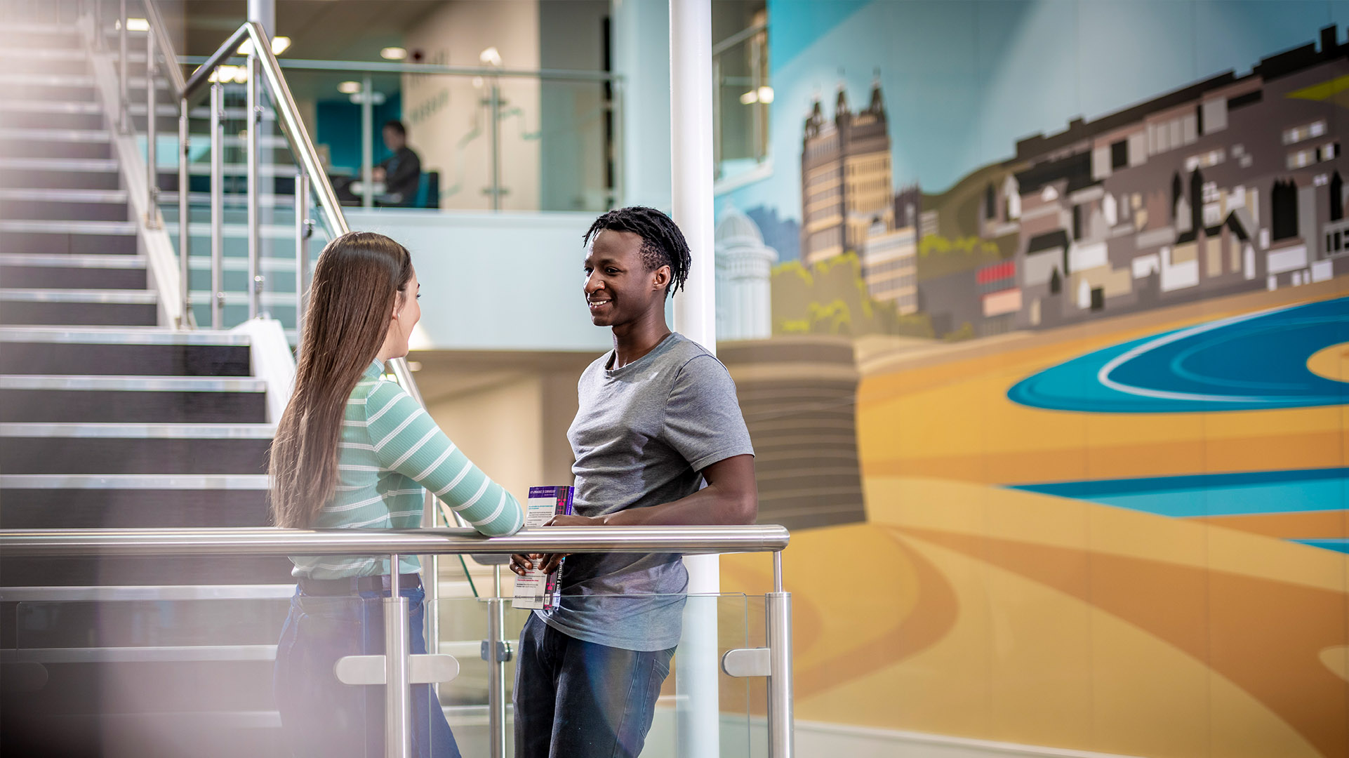 Two students talking on the stairs at CU Scarborough, in front of a mural of Scarborough beach.