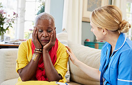 A nurse comforting an older woman sat on a sofa with her head in her hands.